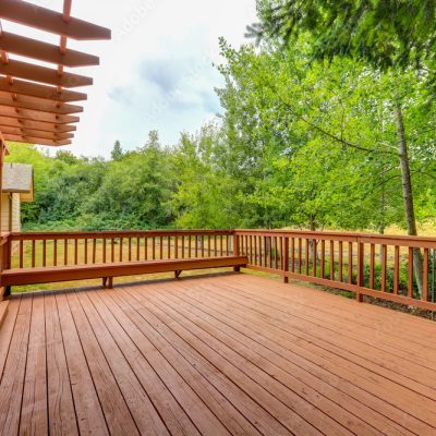 A wooden deck with a pergola and built-in benches overlooks a green, tree-filled backyard. The deck, designed with expert deck building techniques, is attached to a beige house with windows and sits on a grassy area.