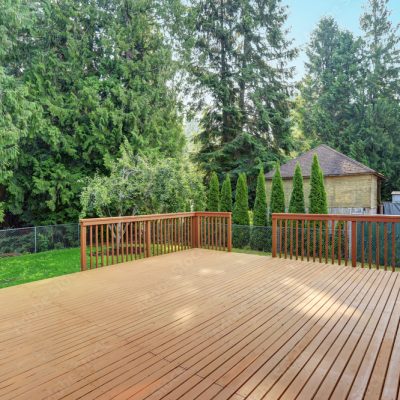 A wooden deck, expertly crafted by a Deck Building and Repairs company, is attached to a house overlooking a green backyard with tall trees and a shed. The sky is clear.