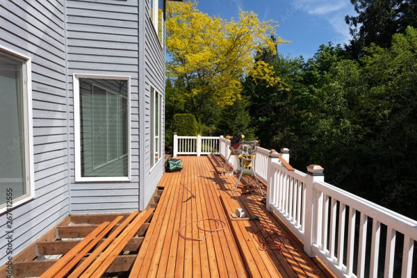 A person is working on building a wooden deck attached to a gray house, focusing on extensions and repairs. Tools and materials are scattered around, while white railings line the edges of the deck. Trees provide a serene backdrop to the ongoing deck building project.