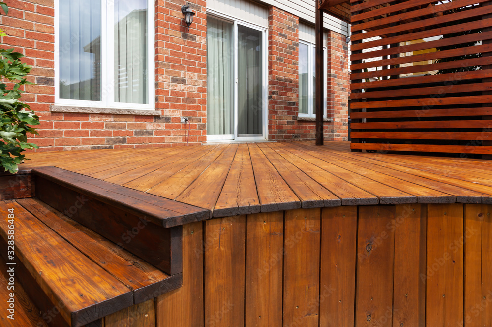 A wooden deck next to a brick house, expertly crafted through deck building, featuring steps on the left and a privacy screen on the right. Large windows and a door in the background seamlessly blend this extension into the home's original design.