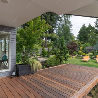 A modern backyard with a wooden deck built by expert deck building and repairs services, attached to the house. It overlooks a landscaped garden with two orange lounge chairs, trees, and a lawn.